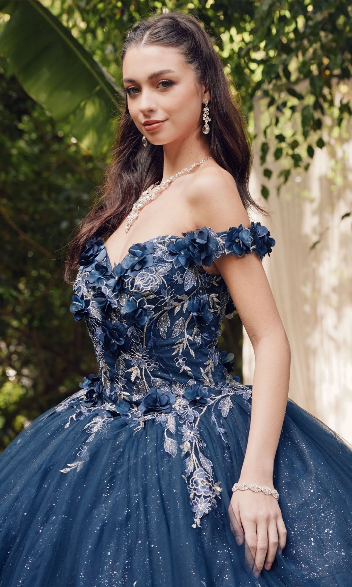 A woman stands outdoors in the Juliet 1447 Off-the-Shoulder Quinceañera Dress, featuring navy blue fabric, beaded embroidery, and glitter details, elegantly accessorized with jewelry.